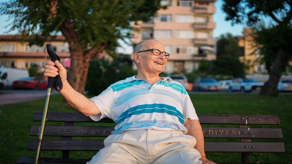 A smiling elderly man sits on a bench holding a walking stick in the park.