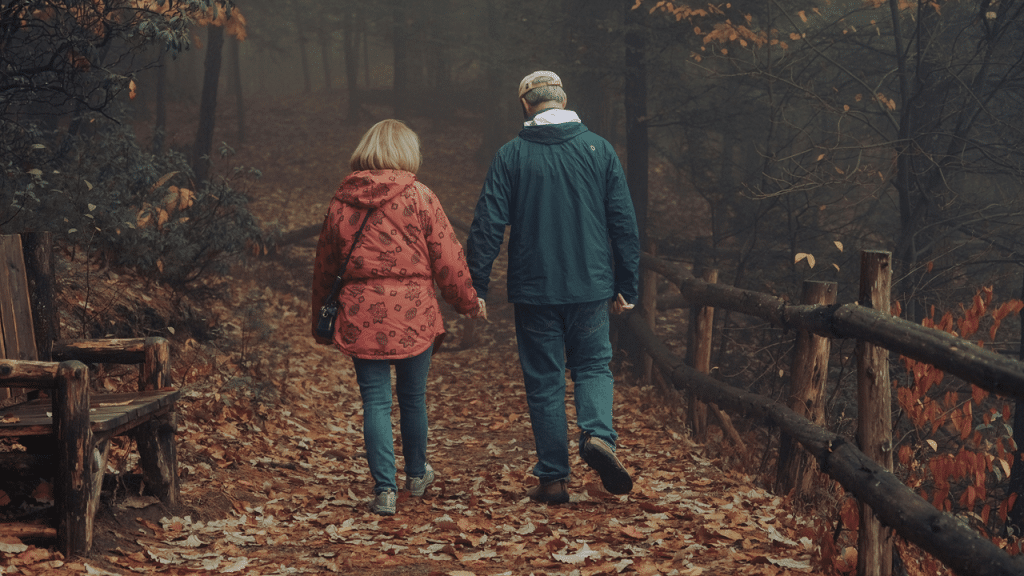 An older couple walking hand in hand through a forest path covered in autumn leaves.