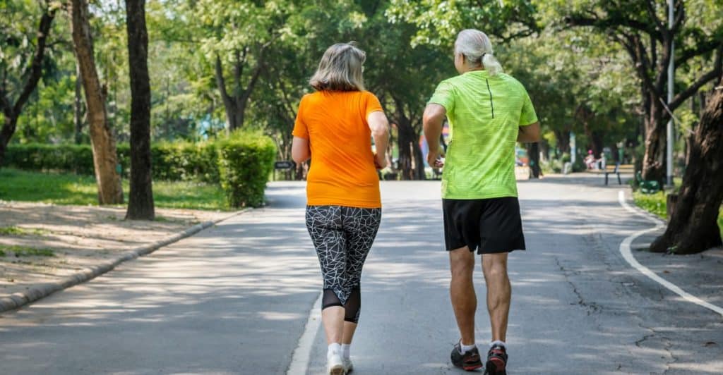A senior couple jogs together down a tree-lined path in the park.