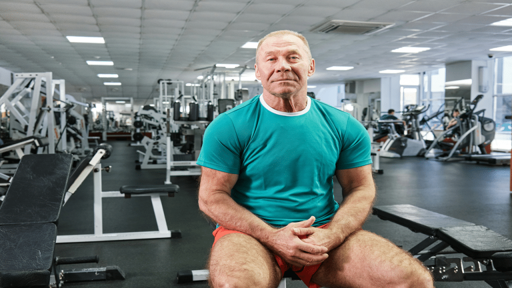 An older man in a teal shirt sitting on a bench in a gym, surrounded by workout equipment. 