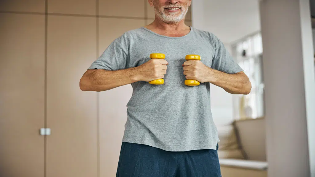 A smiling older man holding two small yellow dumbbells while exercising indoors.