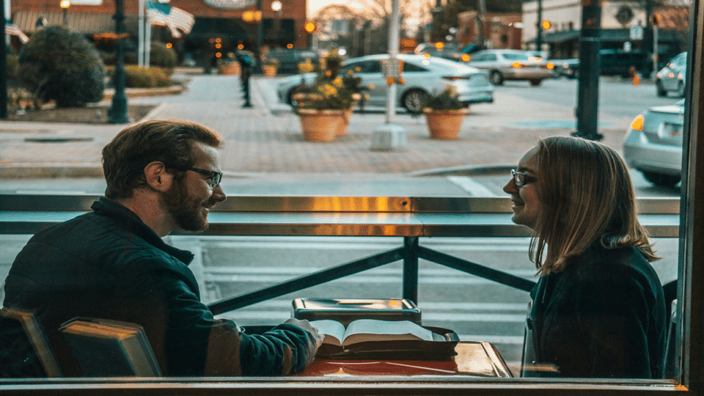 A man and woman smile at each other over an open book in a cozy café, with a street view outside.