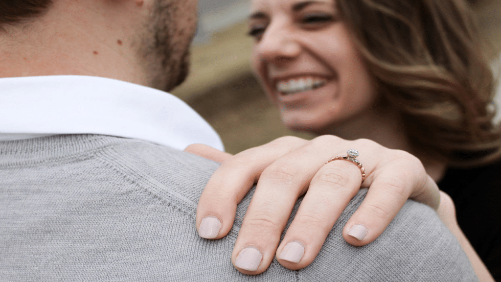 A smiling woman showing off her engagement ring with her hand resting on a man's shoulder.