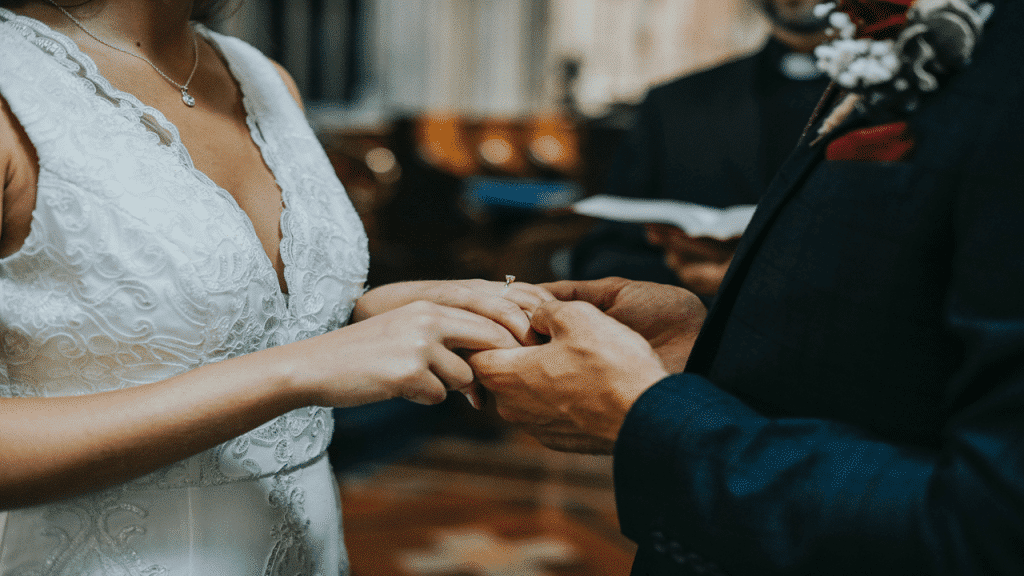 A bride and groom exchanging rings during a wedding ceremony, with a focus on their hands and wedding attire.
