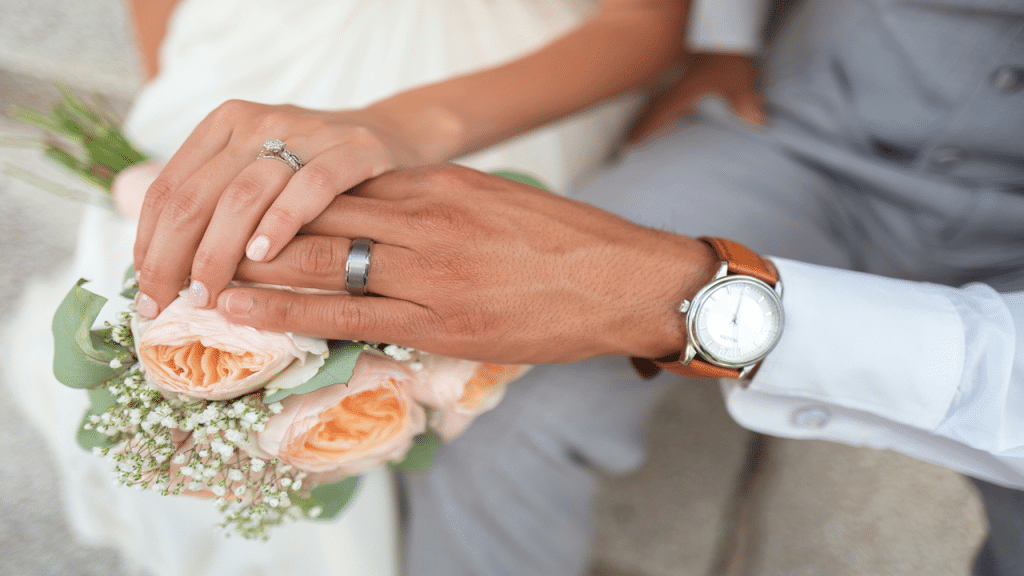 Close-up of a couple’s hands displaying their wedding rings over a peach rose bouquet.
