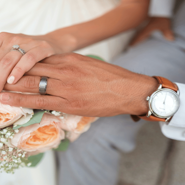 Close-up of a couple’s hands displaying their wedding rings over a peach rose bouquet.