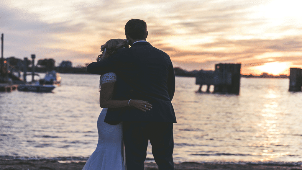 A bride and groom hold each other, facing a tranquil waterfront during a golden sunset.