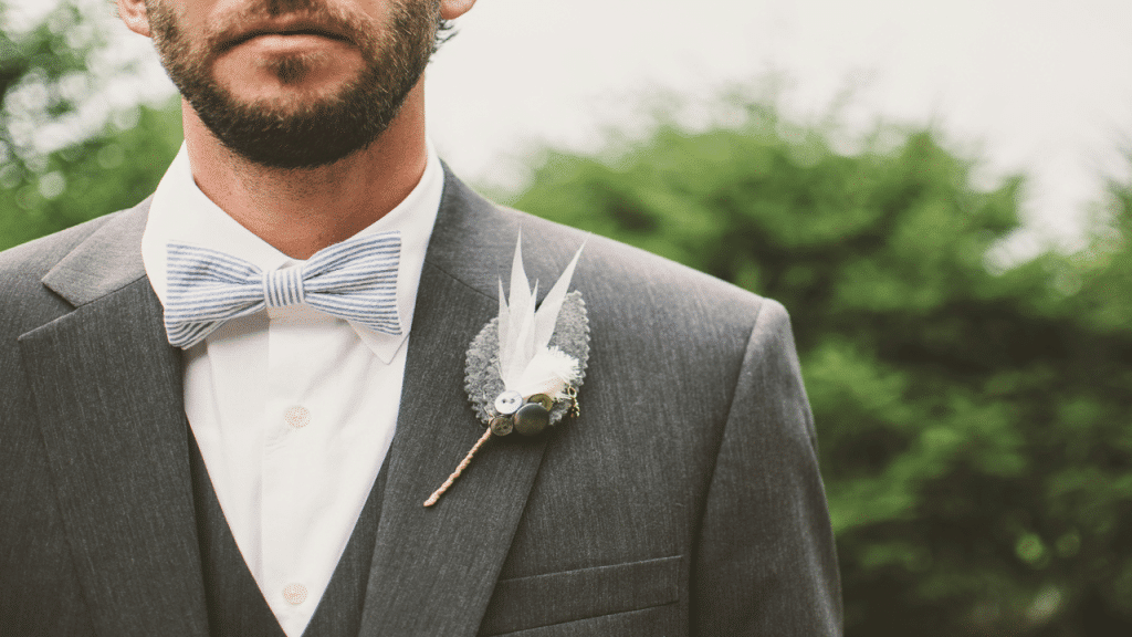 A close-up of a groom in a gray suit and striped bow tie with a decorative boutonniere.