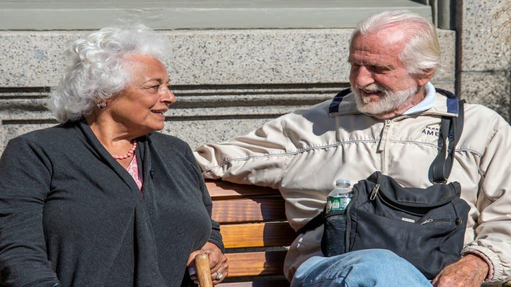 An old couple smiling at each other while sitting on a bench.