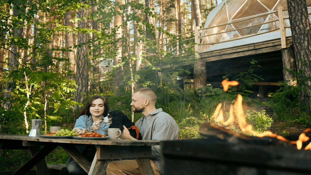 A couple with a dog are eating out in the woods by a fire.