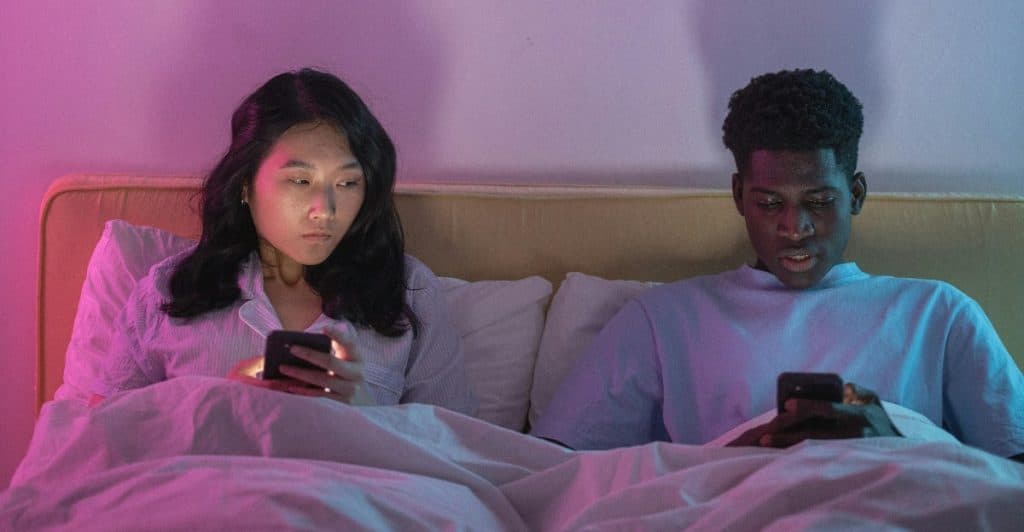A woman looks at her partner scrolling on his phone late at night, ignoring her presence.