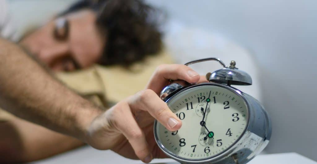 A blurry man sleeps in bed while a hand reaches to stop an alarm clock on a nightstand.