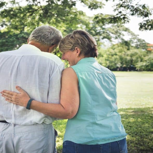 An elderly couple standing underneath a tree and enjoying their time outdoors.