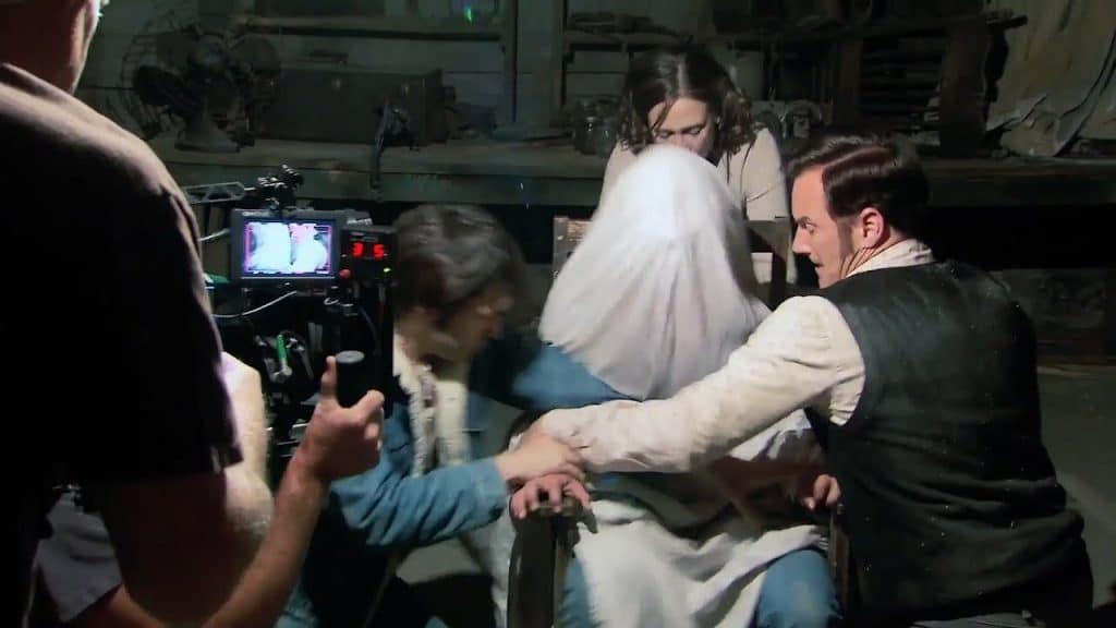 A group of actors subduing another who's covered with a sheet while being filmed by a cameraman.