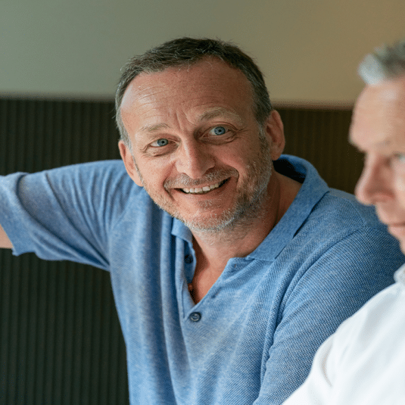 A smiling middle-aged man in a blue shirt gestures while talking with two other men.