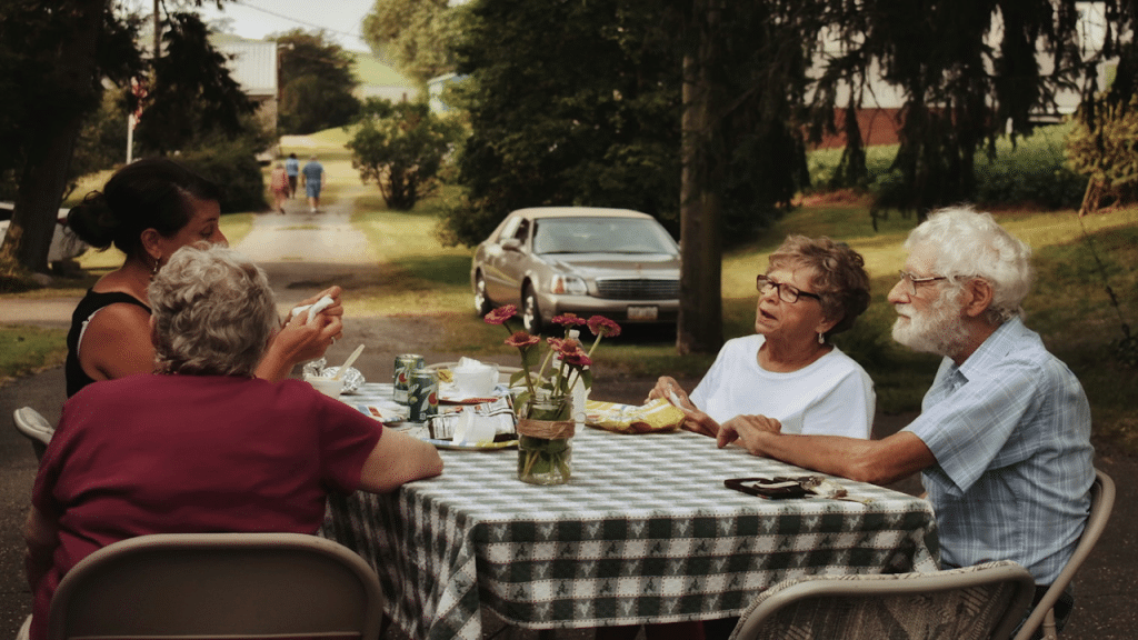 A group of older adults sitting around a checkered table outdoors, sharing a meal and conversation on a quiet driveway lined with trees.
