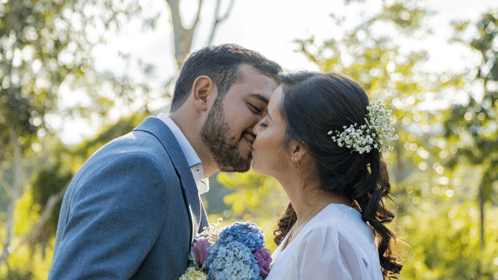 A couple shares a gentle kiss surrounded by greenery, holding a pastel-colored bouquet.