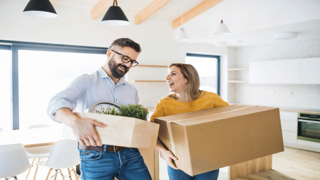 A happy couple carrying moving boxes inside a bright, modern kitchen.