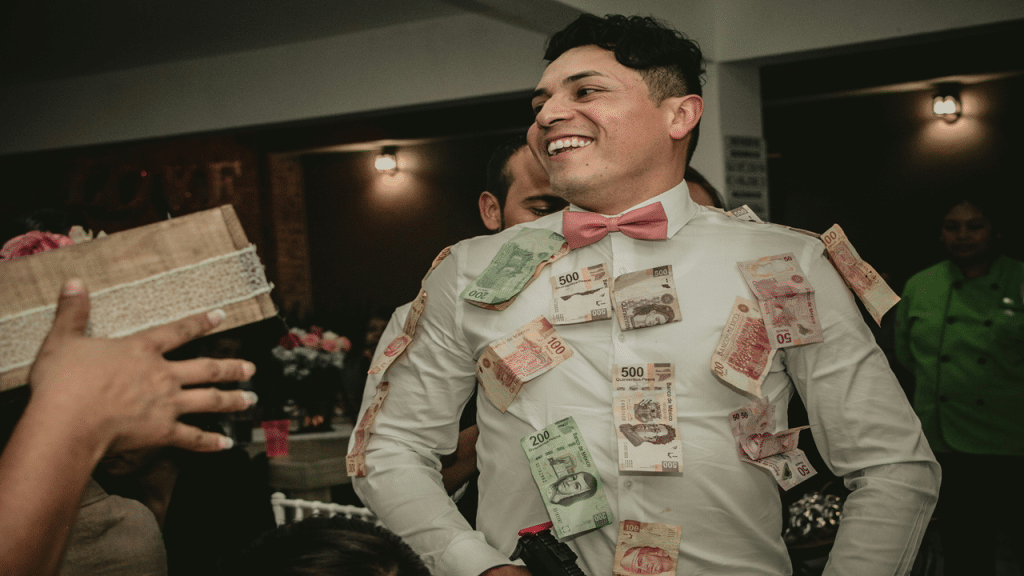 A smiling groom in a white shirt and pink bow tie has various bills pinned to him during a festive celebration.