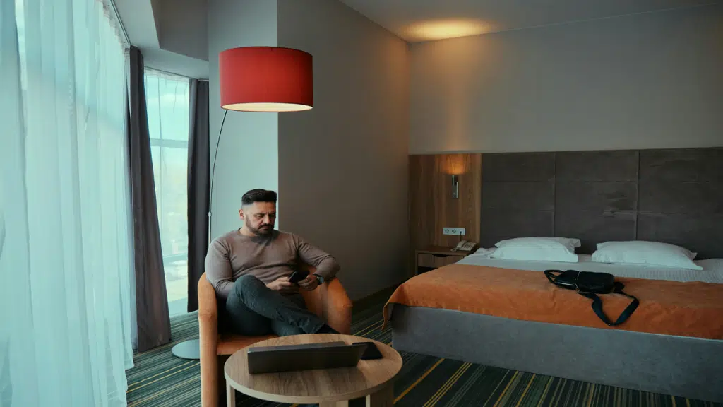 A man sits in a hotel room chair using his phone, with a laptop on the table nearby.