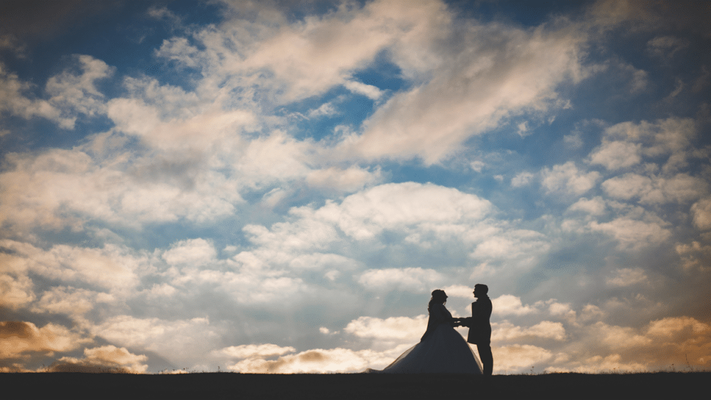 A bride and groom hold hands, silhouetted against a dramatic cloudy sunset sky.