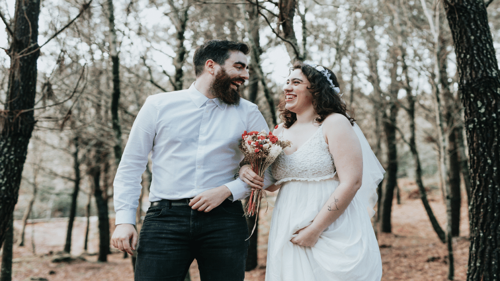A bride and groom joyfully walk through a wooded area, holding a bouquet and smiling at each other.