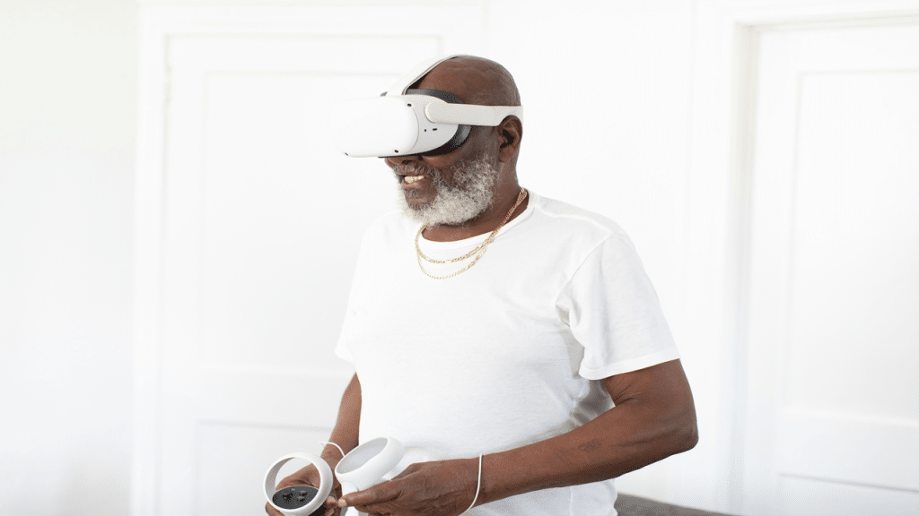 A smiling older man in a white shirt uses a virtual reality headset and controllers indoors.