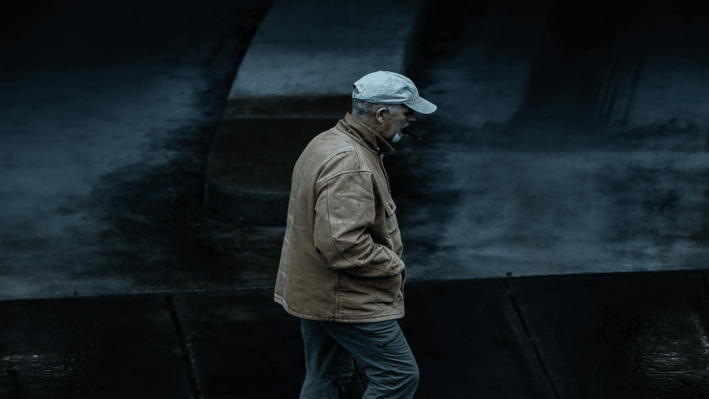A man in a tan jacket and gray cap walks alone on a dark, wet street while smoking a cigar.