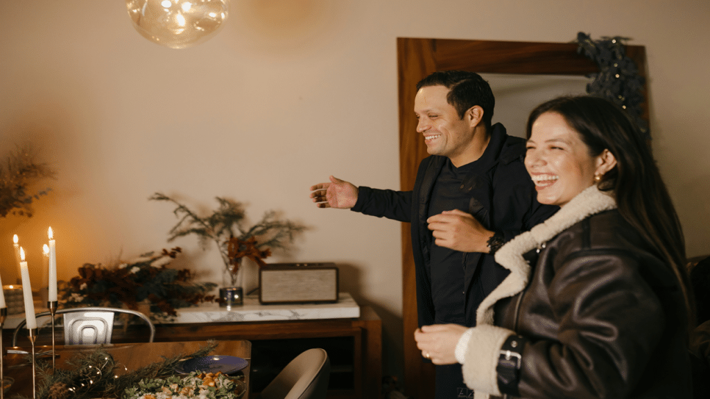 A smiling couple entering a warmly lit dining room decorated for the holidays, with candles and festive greenery on the table.