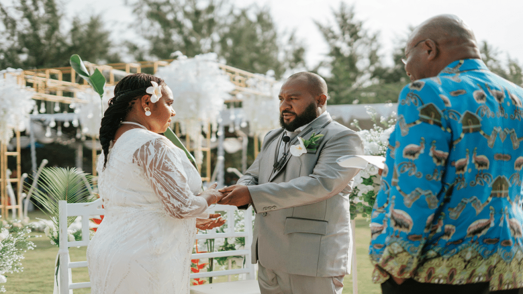 A groom places a ring on the bride's finger during a daytime outdoor wedding with floral decor.