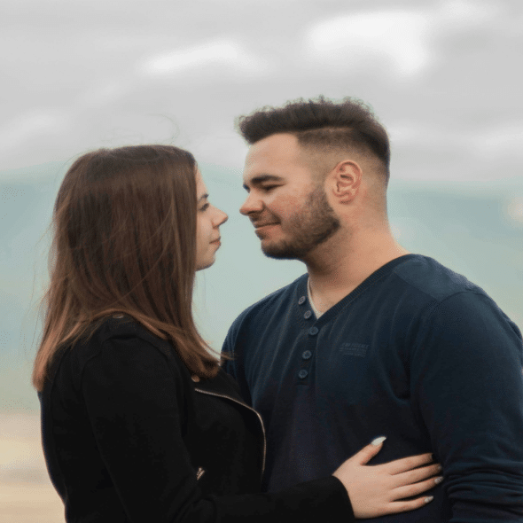 A couple stands close together, gazing into each other’s eyes with a blurred mountain background.