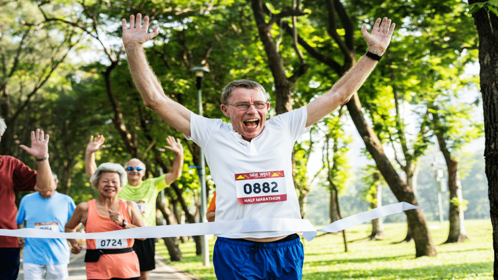 A joyful older man crosses the finish line of a half marathon with arms raised in victory.