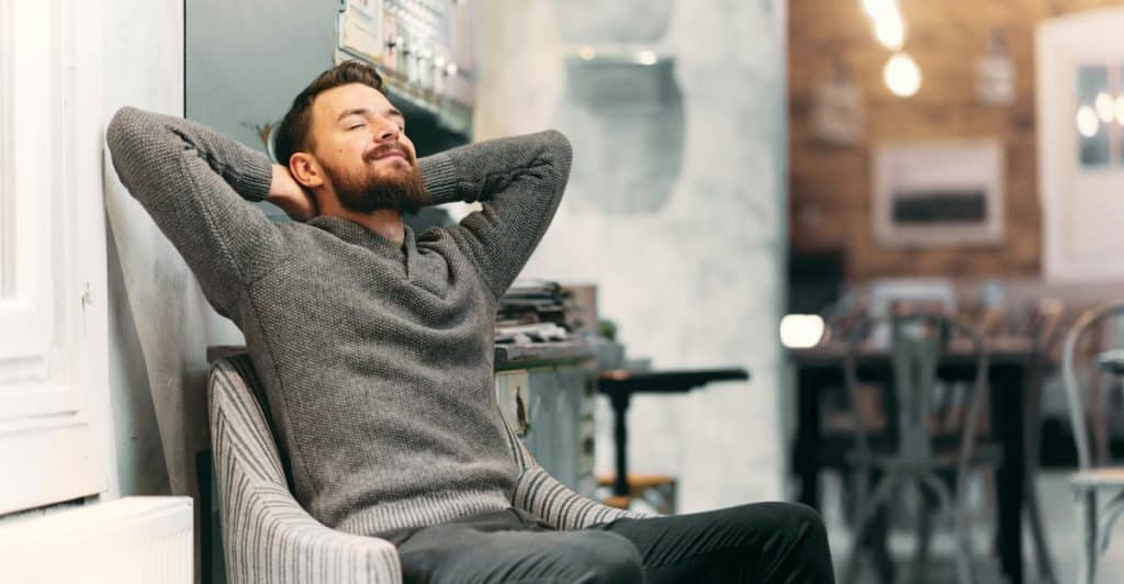 A smiling, bearded man in a gray sweater relaxes in a chair, hands behind his head.