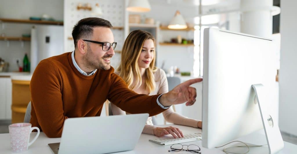 A man in a brown sweater and glasses points at a computer monitor while a woman sits next to him.
