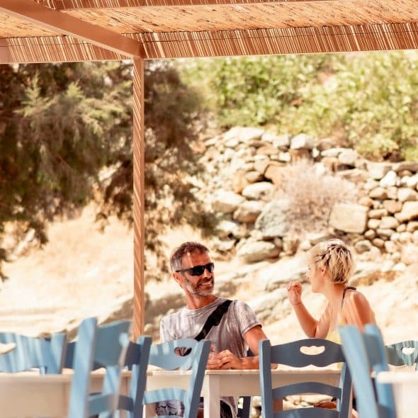 An outdoor café with a man and woman chatting under a bamboo canopy, surrounded by blue chairs and a stone wall.