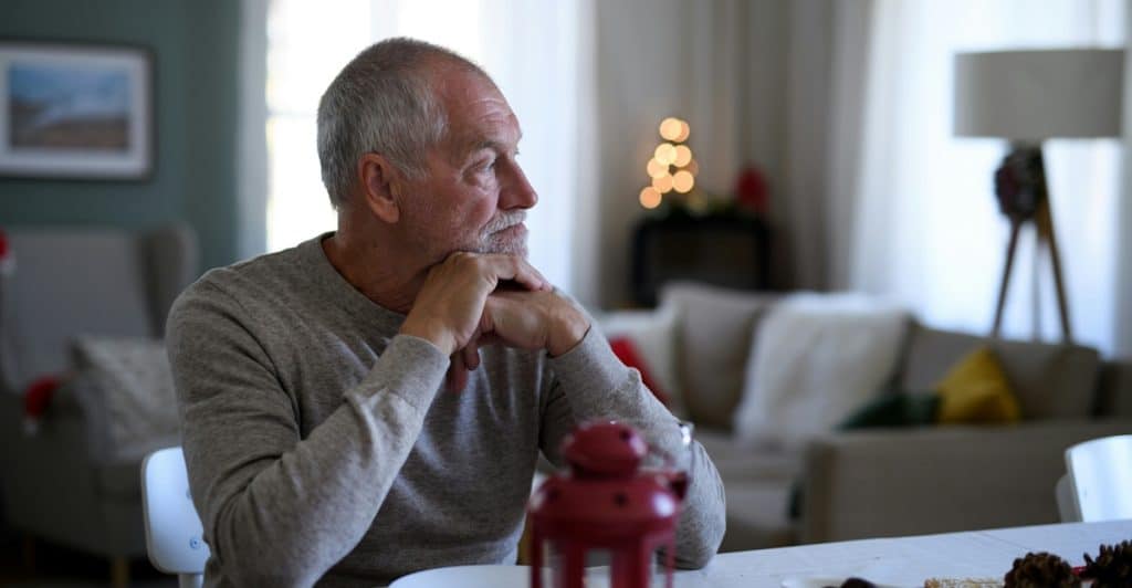 A thoughtful senior man with a beard sits at a dining table, looking to his right.