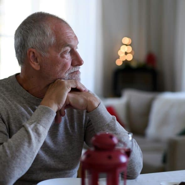 A thoughtful senior man with a beard sits at a dining table, looking to his right.
