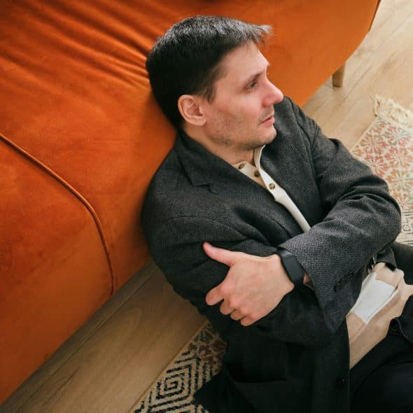A man sits on a rug, leaning against an orange couch, looking to his right.