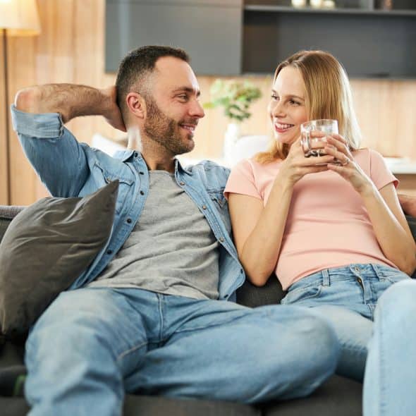 A happy couple smiles at each other while relaxing on a sofa, with the woman holding a glass.