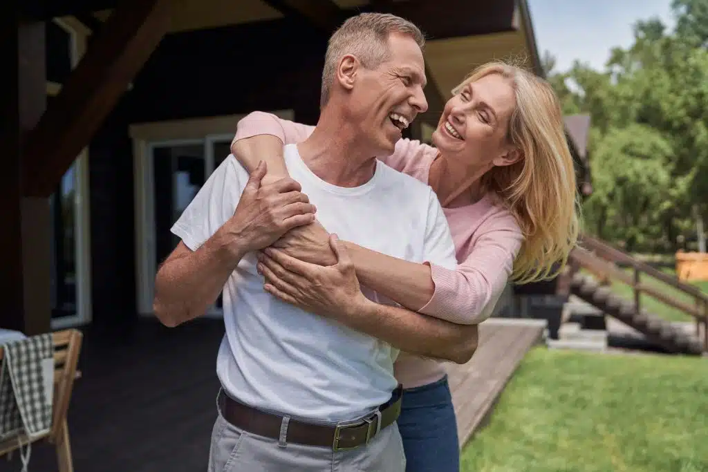 A happy man in a white shirt and a woman in a pink top embrace and laugh outdoors.