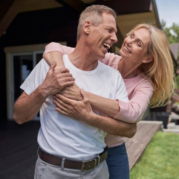 A happy man in a white shirt and a woman in a pink top embrace and laugh outdoors.
