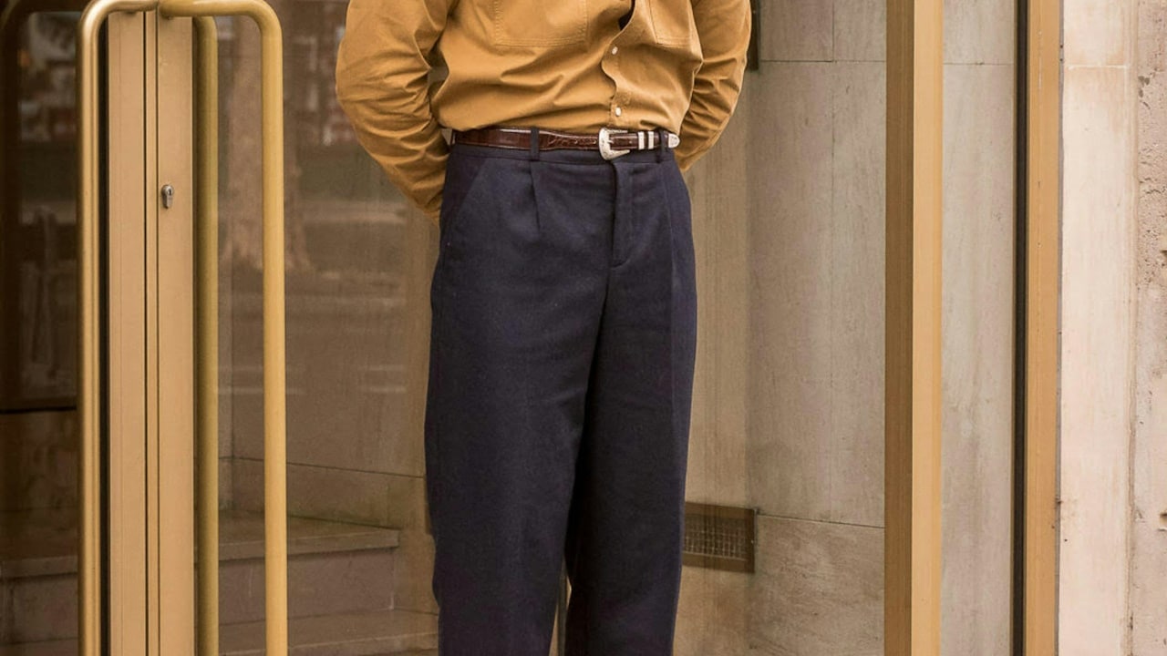 A man wearing a long sleeved shirt over pleated trousers. 