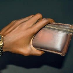 A hand wearing a gold wristwatch holds a worn brown leather wallet with a zipper.