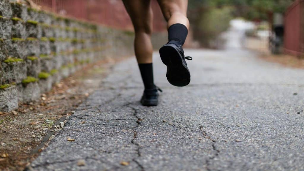 A rear view of a person running on a cracked pavement path, wearing black shoes and high socks.