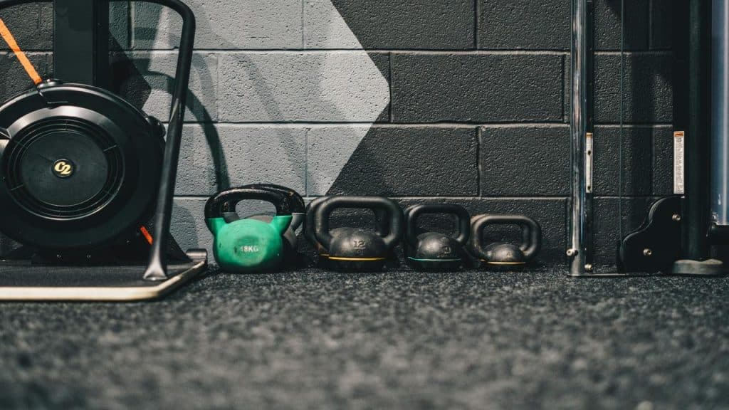 A row of kettlebells lined up on a gym floor next to exercise equipment, with a black and gray painted wall in the background.
