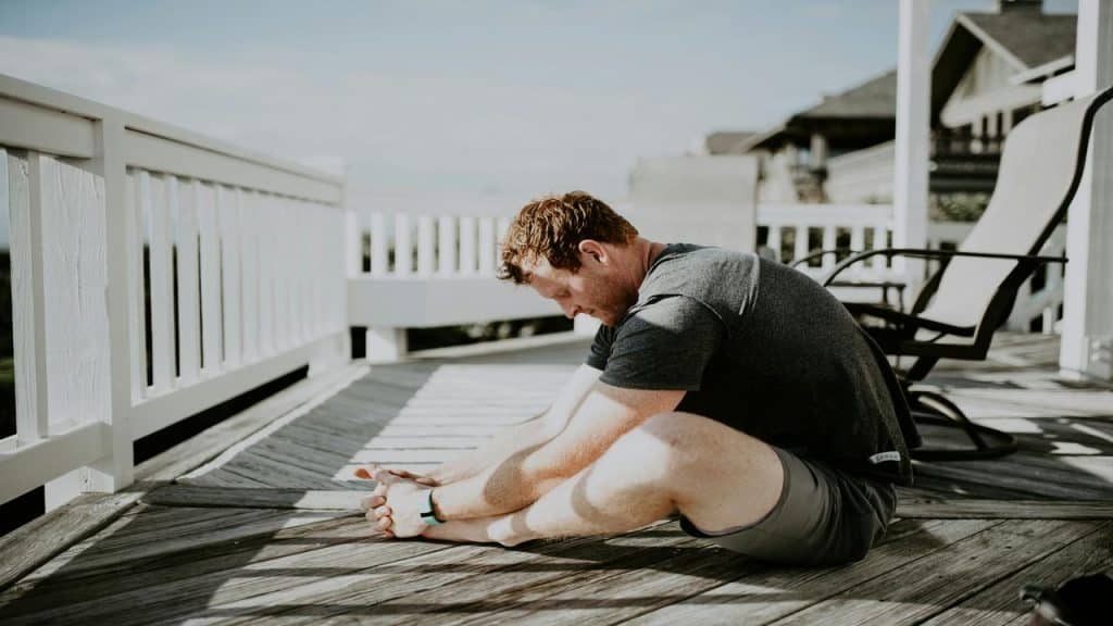 A man sitting on a wooden deck and stretching forward to touch his feet in the sunlight.
