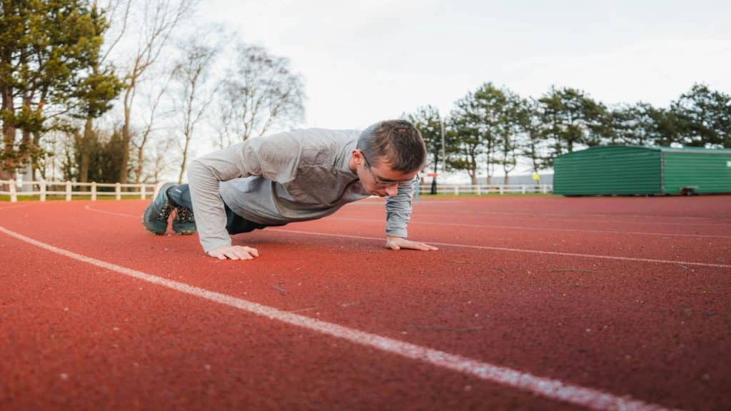 A man in athletic wear doing a push-up on a red outdoor running track surrounded by trees and buildings.