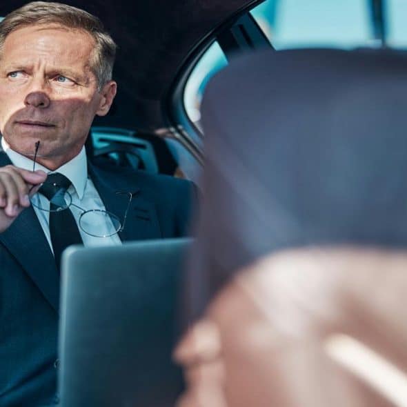 An older businessman in a suit sits in a car, holding glasses and looking out the window.