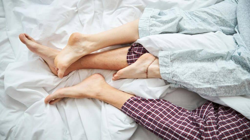A close-up of a couple's intertwined feet in bed, both wearing pajamas, lying on white sheets.