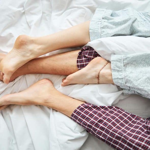 A close-up of a couple's intertwined feet in bed, both wearing pajamas, lying on white sheets.